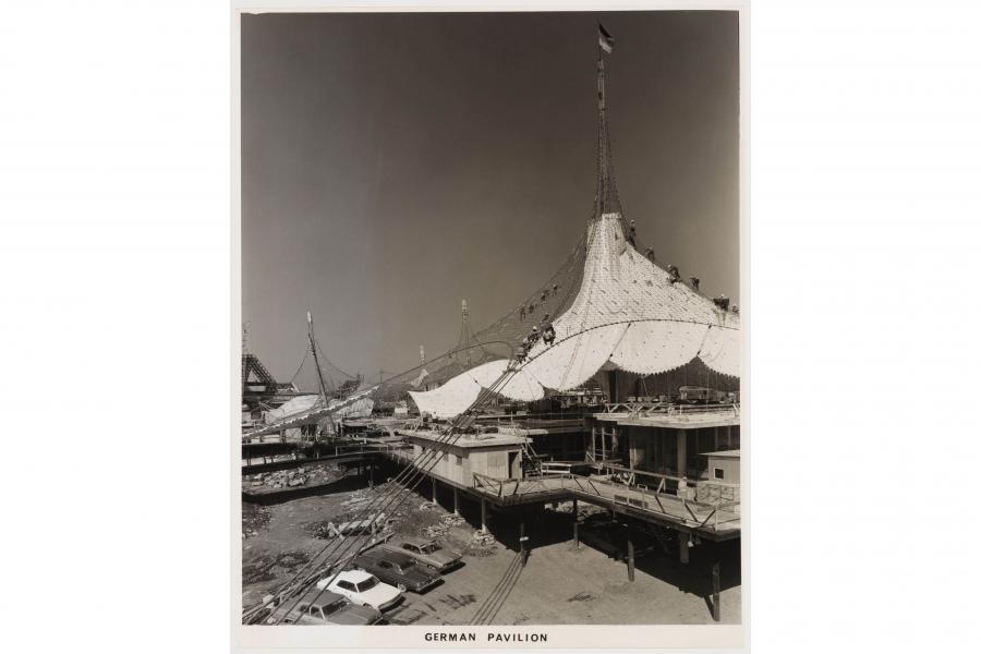 Black-and-white photo of the German Pavilion, showing a sweeping tent-like roof, tall spire, elevated walkways, boats, and parked cars.
