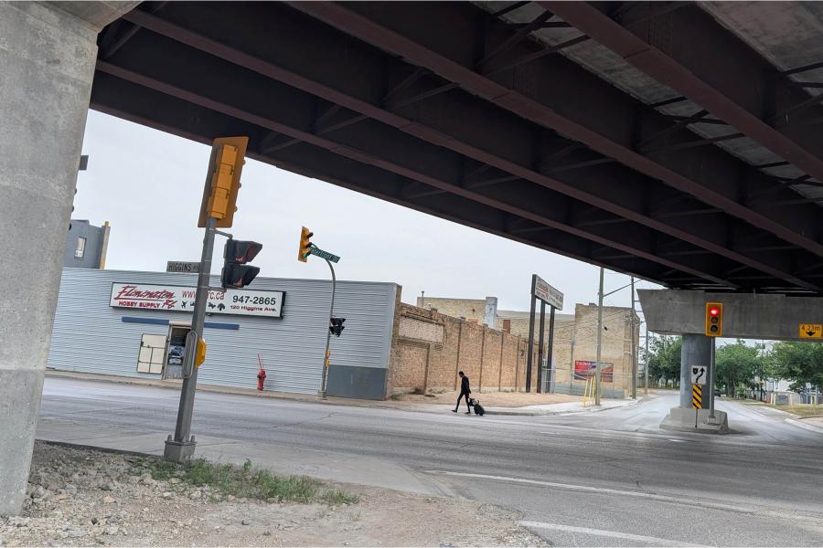 Pedestrian crossing a quiet industrial street beneath a massive steel overpass, with traffic lights, concrete pillars, and muted cloudy skies.