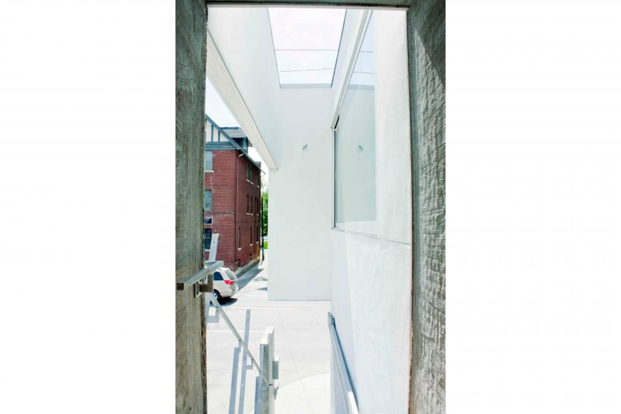 Bright, narrow modern passageway framed by dark walls, with white surfaces, skylights, metal railings, and a red brick building beyond.