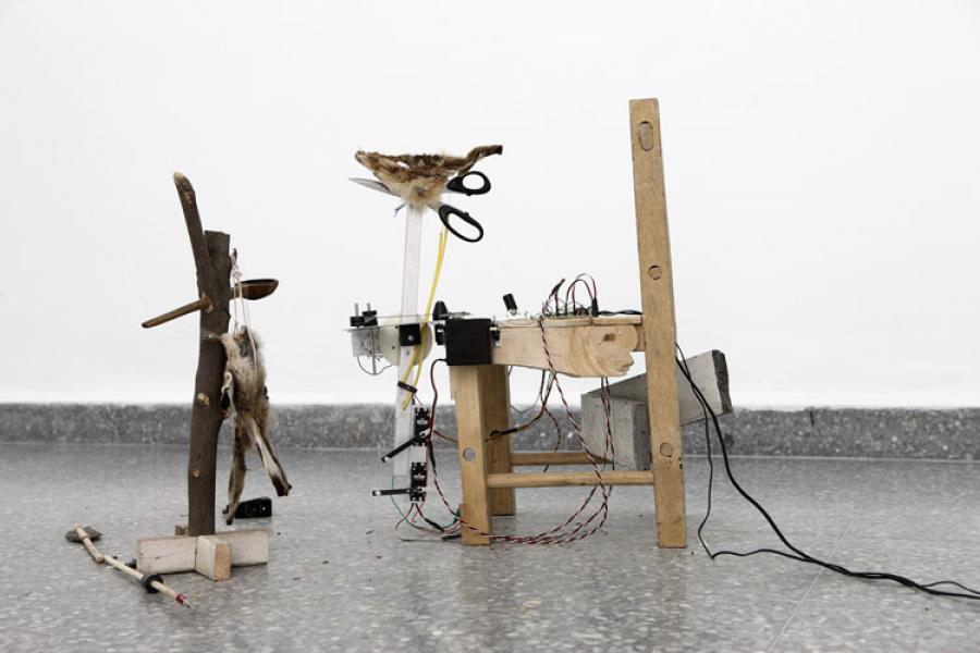 Homemade wooden mechanical contraption with wires, pulleys, and a branch-mounted dried animal form, standing on wet concrete; eerie experimental mood.