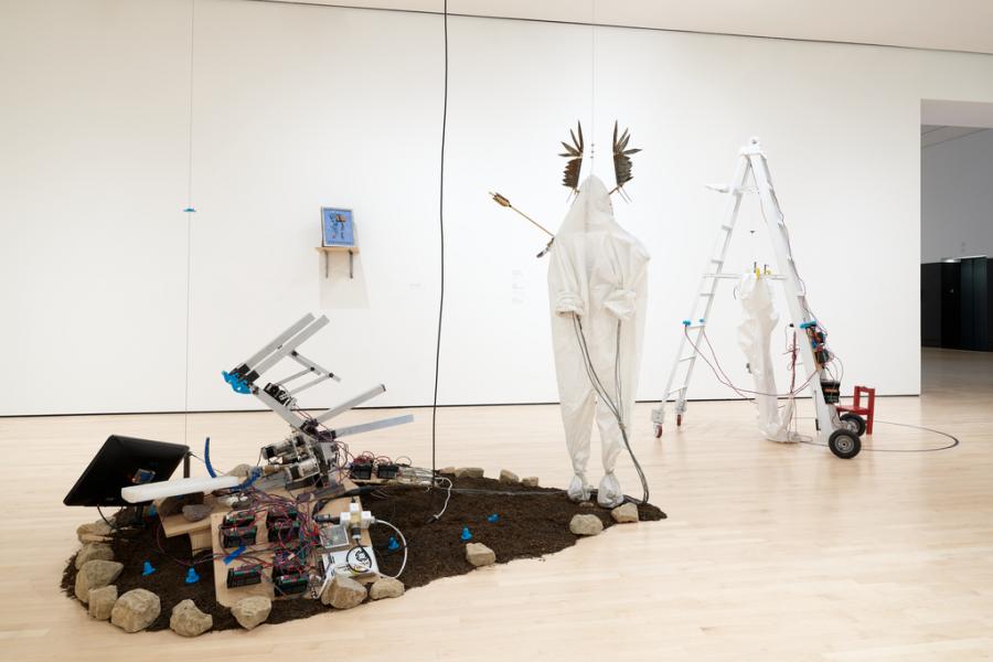 Eerie contemporary art installation in a white gallery: a small rover rests by soil and rocks, beside draped humanoid sculptures, ladders, and cables.