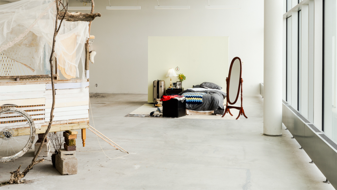 A gallery installation featuring a rustic wooden cart with a weathered bicycle wheel and draped fabric on the left, transitioning to a cozy domestic scene on the right with a bed, mirror, and bedside table in front of a soft yellow backdrop.