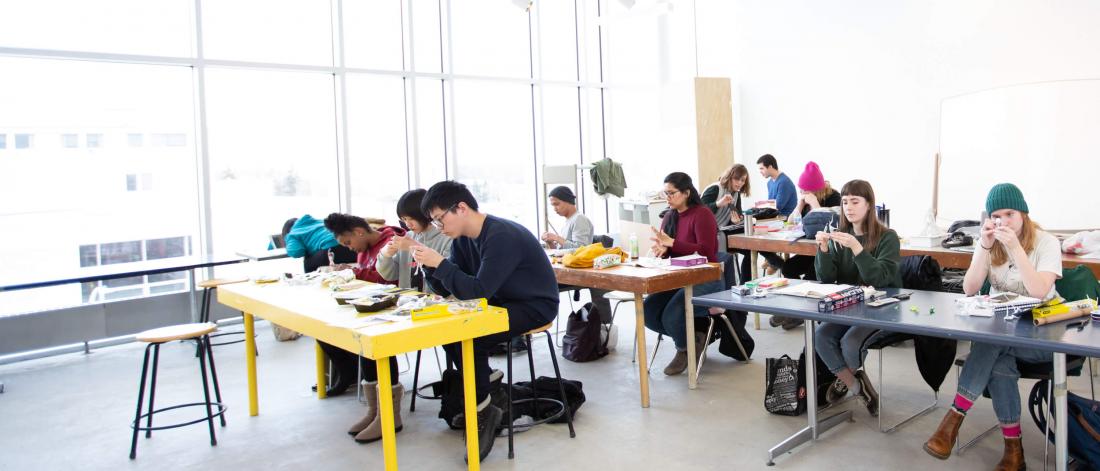 A class of first year school of art students work in a studio at long narrow tables.
