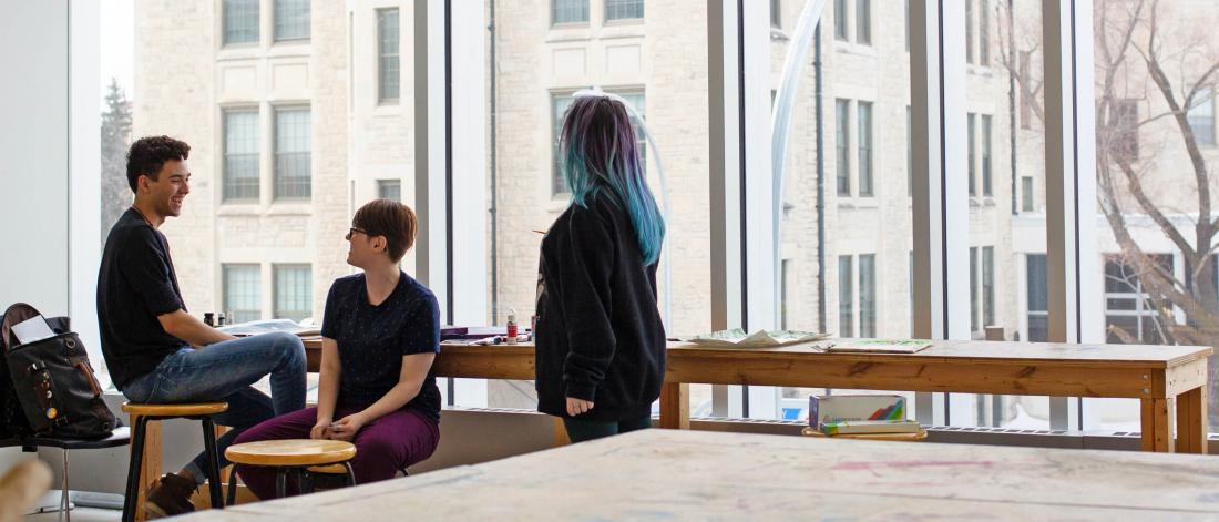 Three School of Art students sit at a long narrow table in front of a window. 