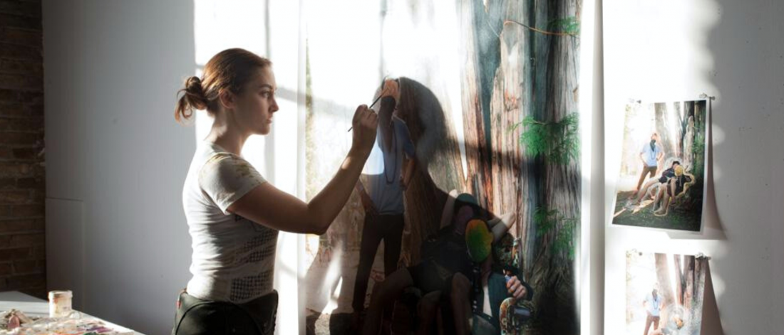 Sarah Anne Johnson paints a large painting, hanging on a wall, in a sun filled studio