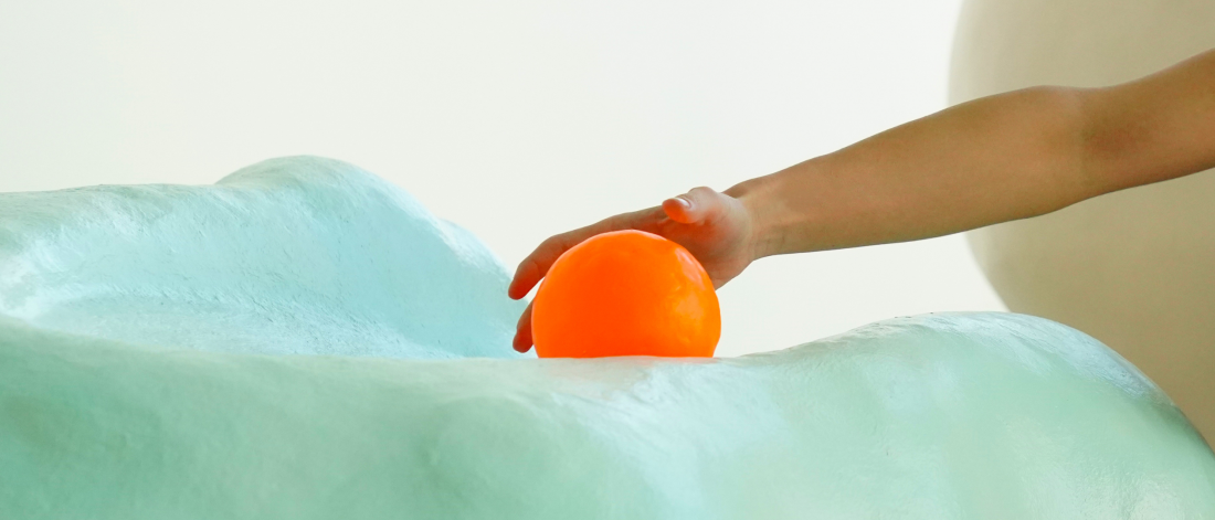 A hand reaches for a a bright orange sphere resting on a light blue/green sculpture.