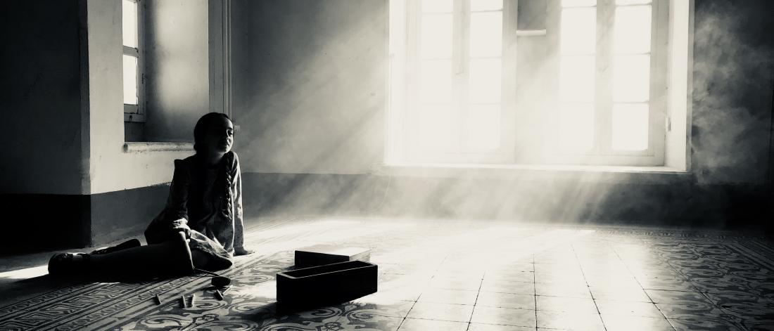 Black and white photograph of light cascading through windows into a figure sitting in am empty room