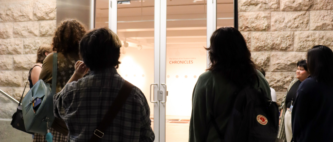 A group of people stand outside the entrance of the School of Art Student Gallery. The sign above the glass doors reads "School of Art Student Gallery."