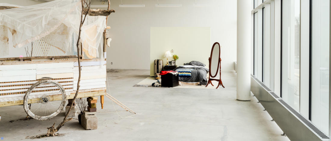 A gallery installation featuring a rustic wooden cart with a weathered bicycle wheel and draped fabric on the left, transitioning to a cozy domestic scene on the right with a bed, mirror, and bedside table in front of a soft yellow backdrop.