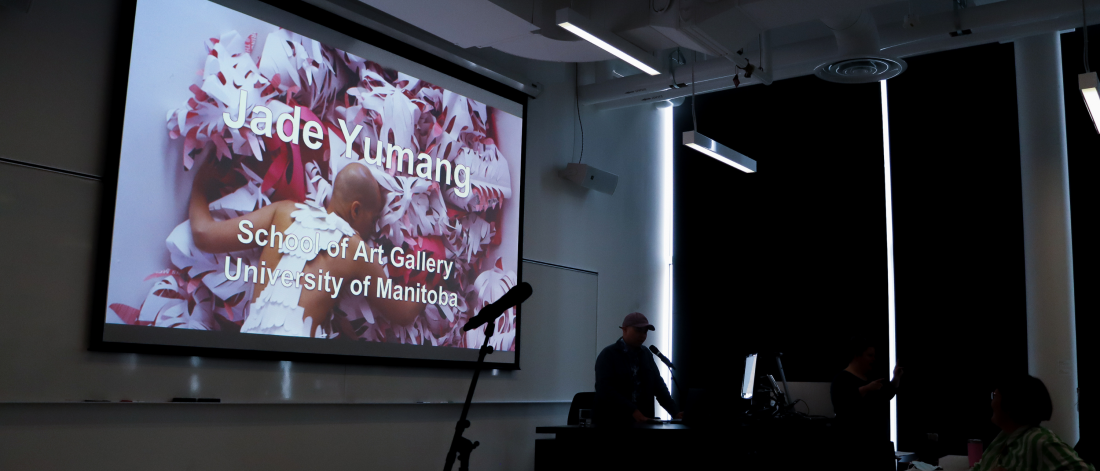 An artist gives a talk in a darkened lecture room with a projected slide reading “Jade Yumang, School of Art Gallery, University of Manitoba.”
