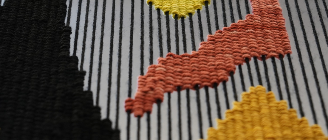 A close-up of woven textile work with geometric patterns in black, orange, yellow, and red.