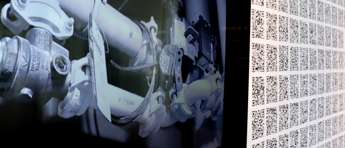 Video still showing complex plumbing and machinery in grayscale on a screen beside the QR code installation.