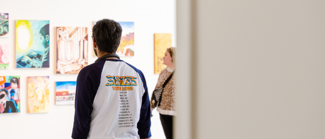 A person wearing a blue-and-white “Exies Tour 2022” shirt stands in a bright gallery space, looking at a wall of colourful paintings. Another visitor in a patterned top is slightly blurred in the background. The foreground is partially framed by an out-of-focus white wall on the right.