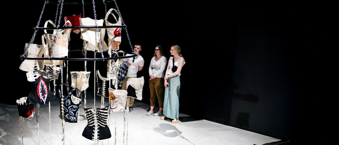 Visitors stand in a gallery observing a suspended circular installation of hanging textile and corset-like forms, arranged on chains in a dimly lit exhibition space.