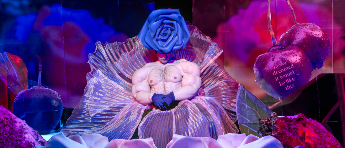 A staged portrait of a performer seated inside an iridescent flower-like form, wearing exaggerated muscular prosthetics and a large blue rose headpiece, surrounded by glowing pink and blue floral imagery and oversized cherries.