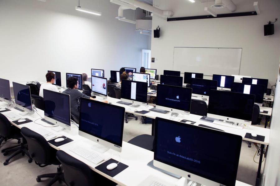 Rows of iMac desktop computers on desks with students sitting at various work stations.