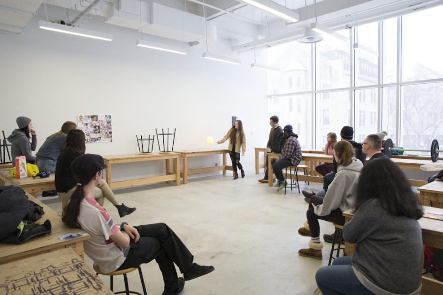Students sit in a circle around a prof for a presentation in a bright window lit ARTlab studio