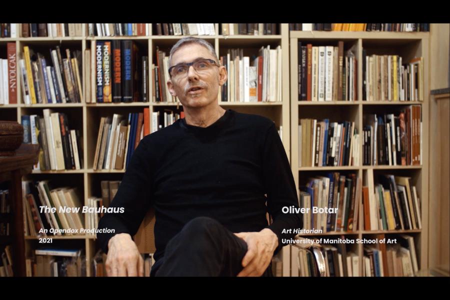 Botar sits in a chair in front of a bookcase full of books, wearing a black sweater.