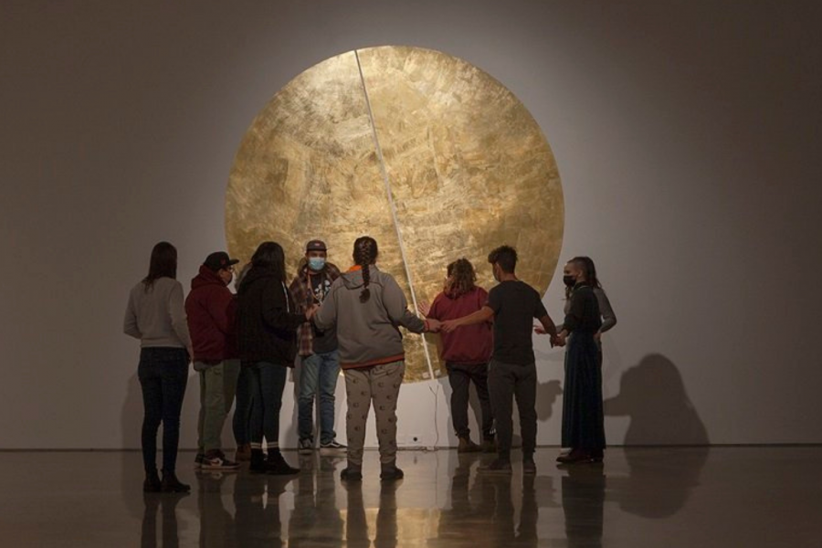 A group of students gather, staring away from camera at a gold metallic circle on the gallery wall.