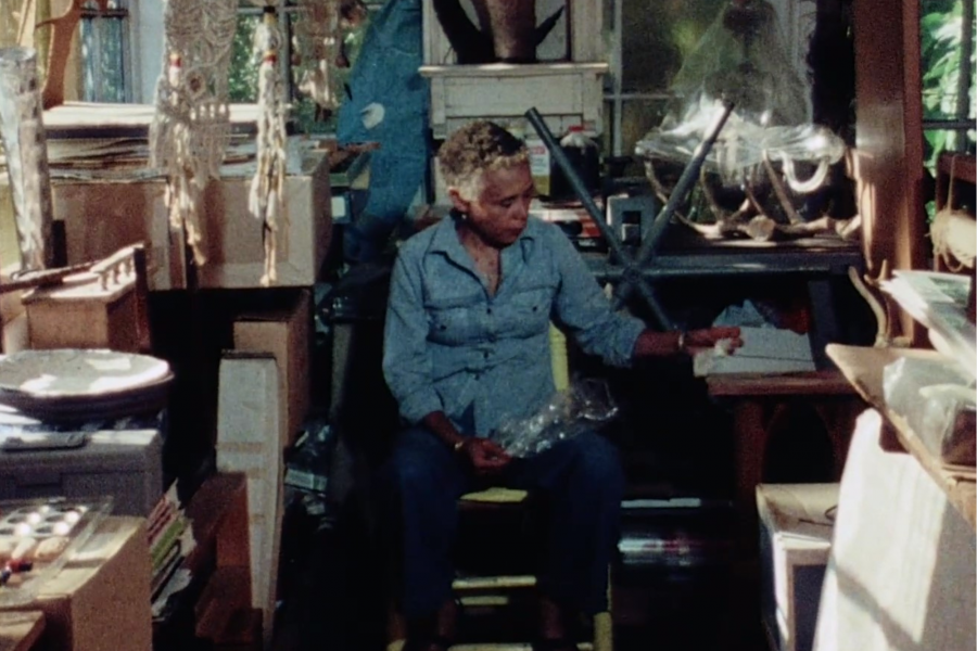 Betye Saar sits in her studio wearing a blue collared shirt, looking down.