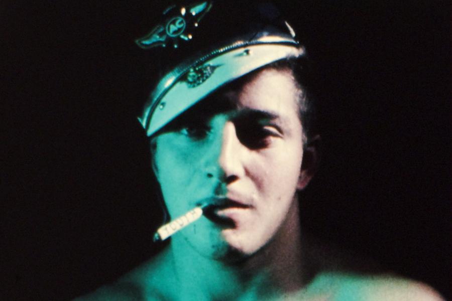 A mans face, wearing a leather hat, cigarette hanging from his mouth. A bluebell light shines on his face.