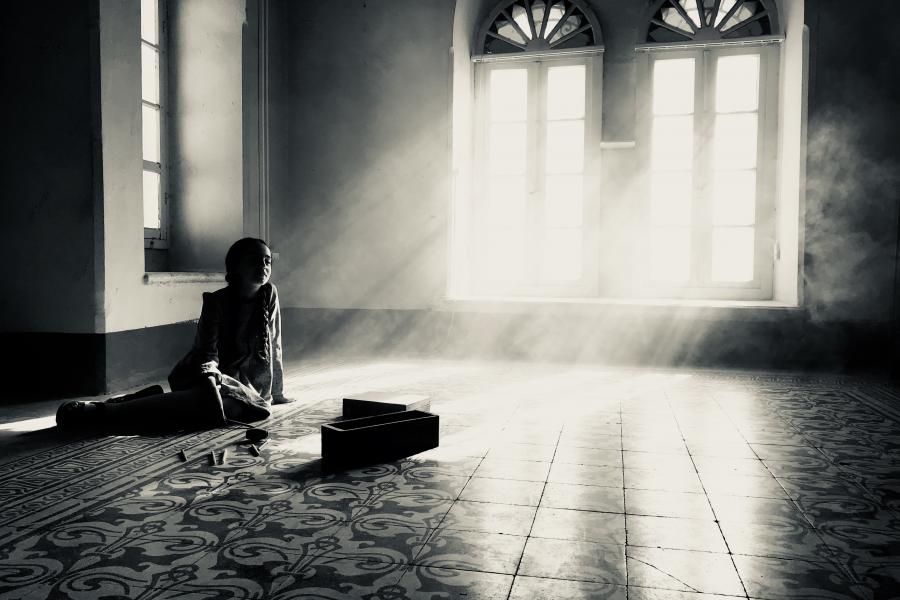 Black and white photograph of light cascading through windows into a figure sitting in am empty room