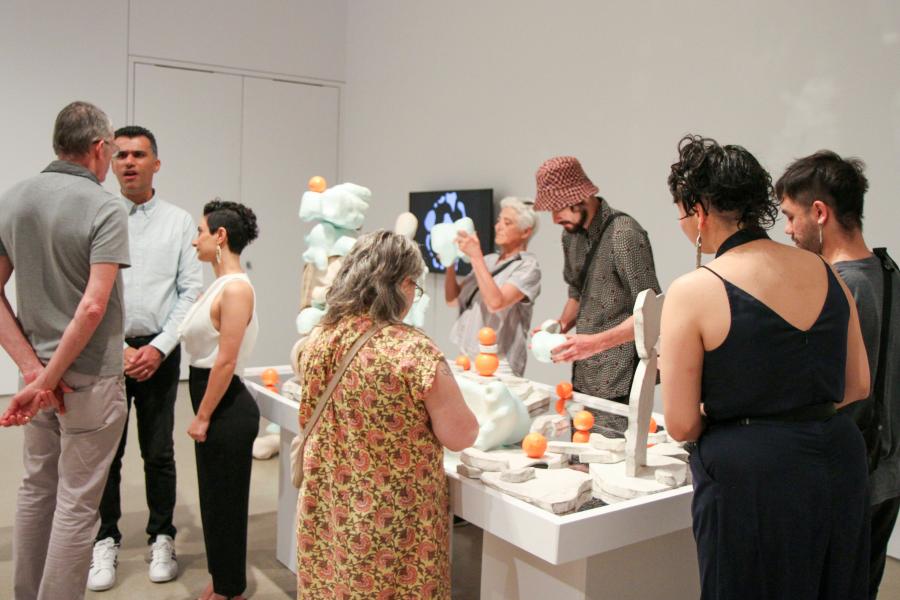 A group a people gather around a white table touch and moving around an art piece of blue and white sculptural forms.