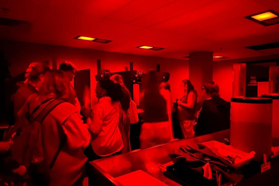 A group of people in a red-lit darkroom, observing photographic equipment and processes.