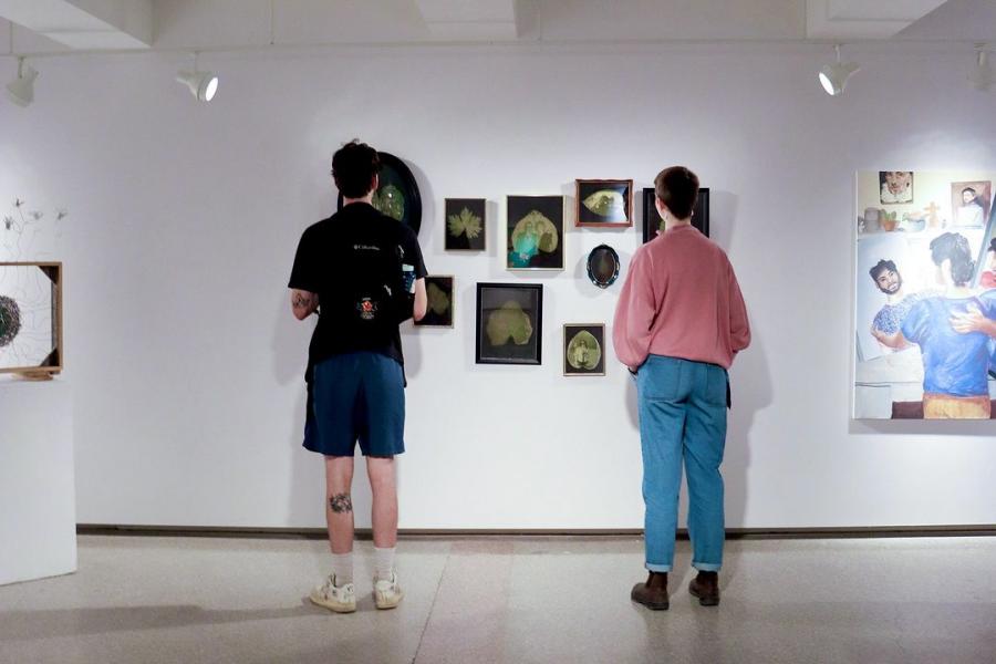 Two visitors view an art display with multiple framed works on a gallery wall.