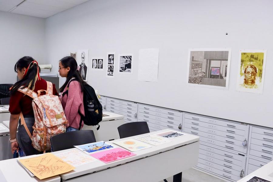 Two students examine printed artworks and posters displayed on tables and walls in a classroom setting.