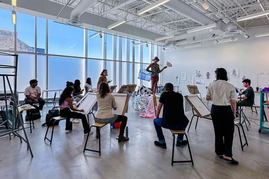 Students participate in a life drawing class, sketching a model posing with props in a bright studio.