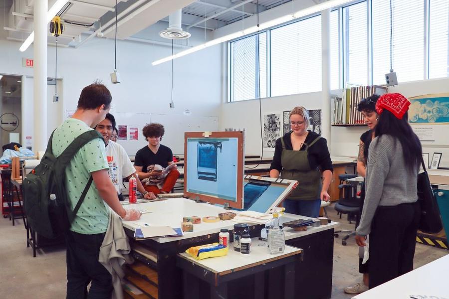 A group of students gather around a table for a printmaking demonstration, engaging with tools and materials.
