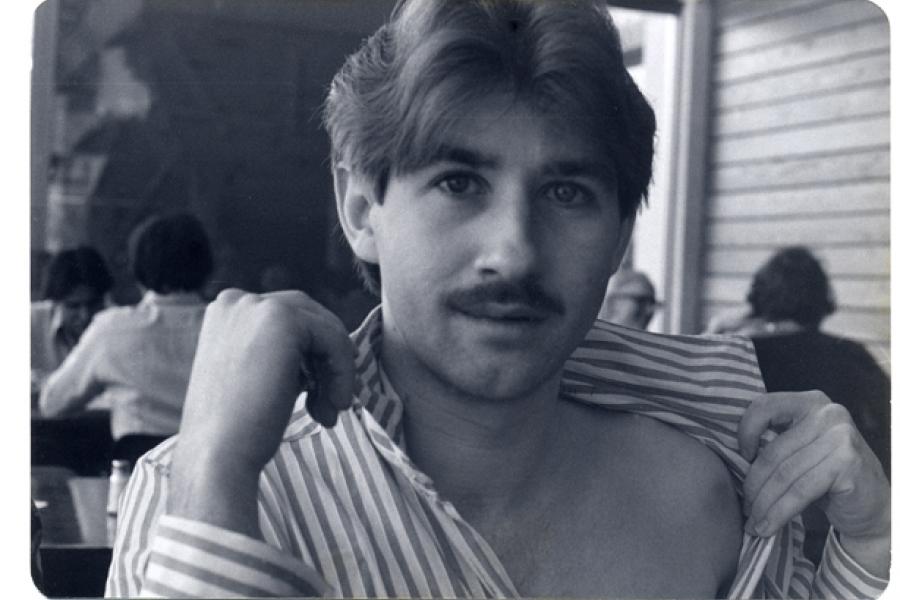  A black and white photograph of a young man with a mustache, gazing directly at the camera while pulling open his striped shirt to reveal part of his chest, with people sitting in the background.
