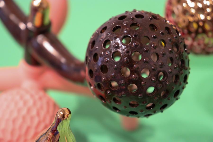 A dark, perforated ceramic sphere with a glossy finish, resting among other vibrant sculptural elements on a green table.