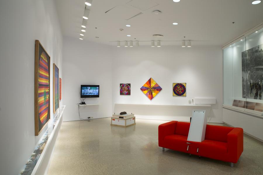 A vibrant gallery installation featuring colorful geometric paintings, a video monitor, and a bright red couch with an interactive panel.
