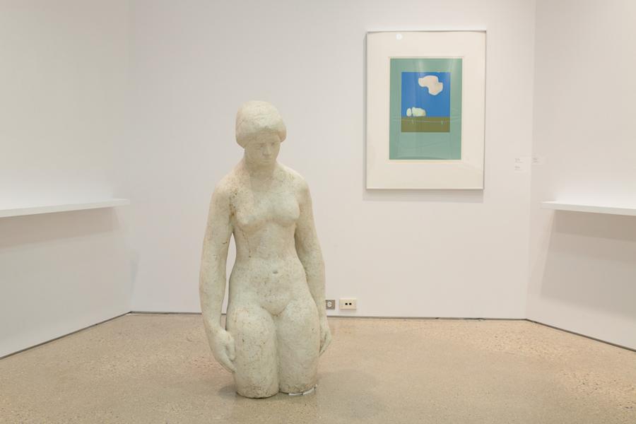 A seated sculpture in a minimalist gallery setting, positioned near a framed artwork depicting a bright blue sky and green landscape.