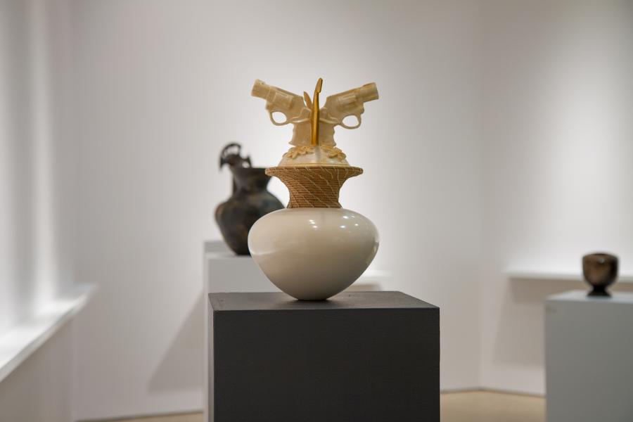 A ceramic vase with intricately crafted revolvers at the top, displayed on a pedestal in a gallery setting.