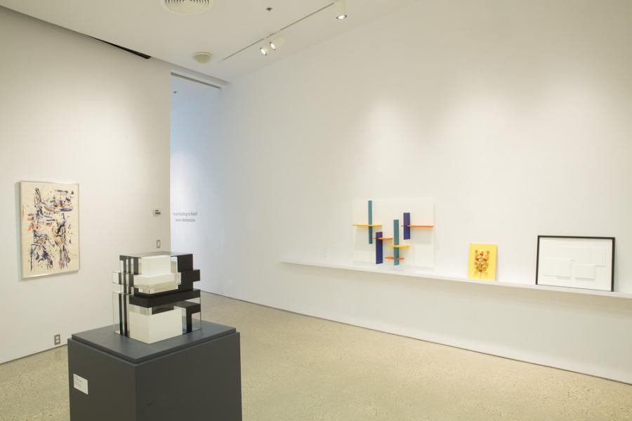 Gallery view showcasing a mix of sculptures and paintings, including abstract works on a shelf and a black and white cubic sculpture in the foreground.