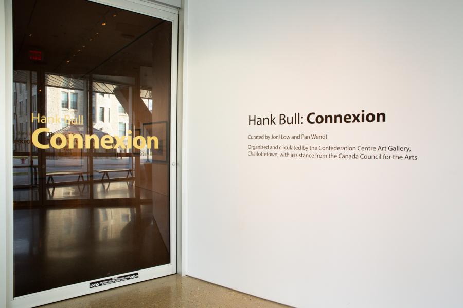 Entrance to the Hank Bull: Connexion exhibition, featuring text on a glass door and wall with curatorial credits.