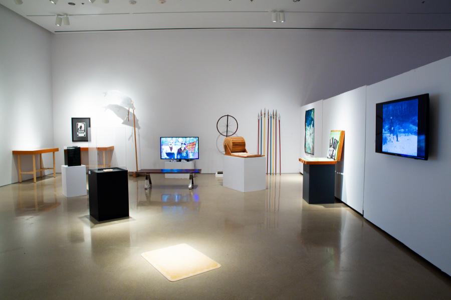 A gallery installation with multimedia works, including a TV screen, colourful spears, and sculptural pieces.