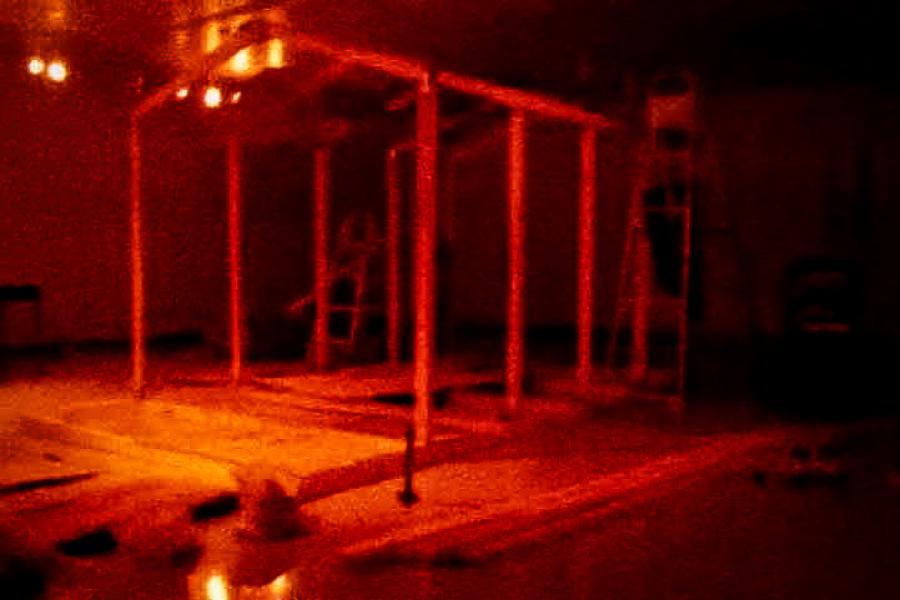 A dimly lit gallery space with an unfinished wooden frame structure resembling the skeletal form of a small house. Ladders and scattered building materials surround the structure, suggesting an in-progress installation.