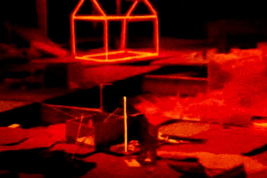 A closer view of the installation, featuring a glowing red neon house frame positioned above a collection of miniature models and scattered materials. The neon light contrasts sharply with the dark surroundings.