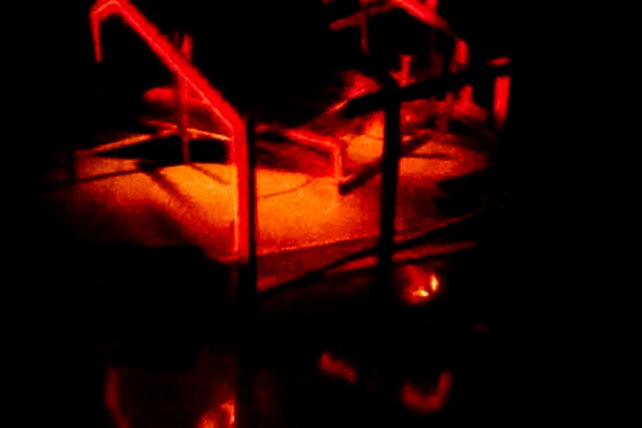 A fragmented wooden framework illuminated by red neon tubing, giving the impression of a partially collapsed or deconstructed structure. Shadows and reflections on the floor add to the atmosphere of impermanence.