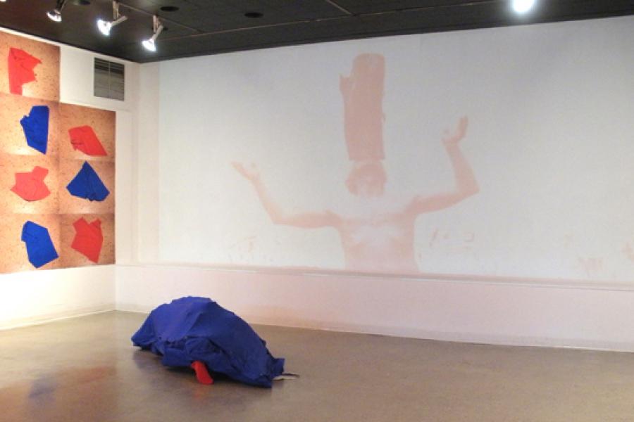 An art installation featuring a large-scale video projection of a shirtless person with a wooden object on their head, a fabric-covered object on the floor, and a wall display of red and blue geometric pieces.