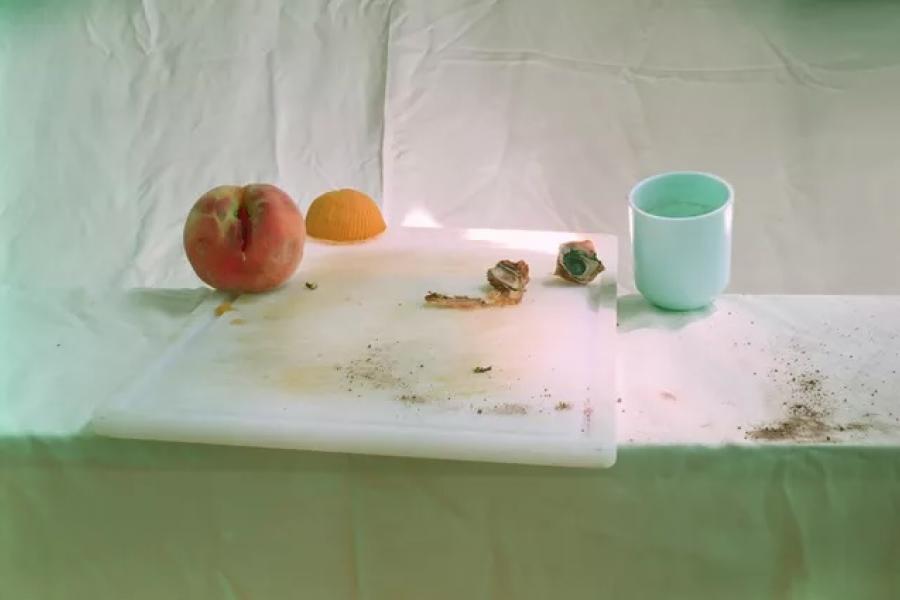A minimalist still life photograph featuring a white table with a cutting board holding a peach, an orange peel, a few scraps, and debris. A plain white mug sits nearby, with faint greenish lighting casting subtle shadows on the white fabric background, evoking a quiet, contemplative atmosphere.