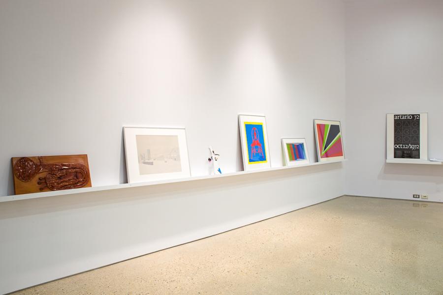 A gallery wall with a long white shelf displaying framed artworks. From left to right: a sculptural piece resembling a musical instrument, a muted urban sketch, a colourful graphic print, a minimalist geometric artwork, and a black-and-white poster reading "artario 72 oct12/1972."