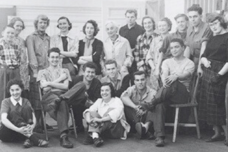 A black and white vintage photograph of a large group of young men and women posing together in a casual setting, some sitting on chairs or the floor and others standing, dressed in mid-20th-century attire.