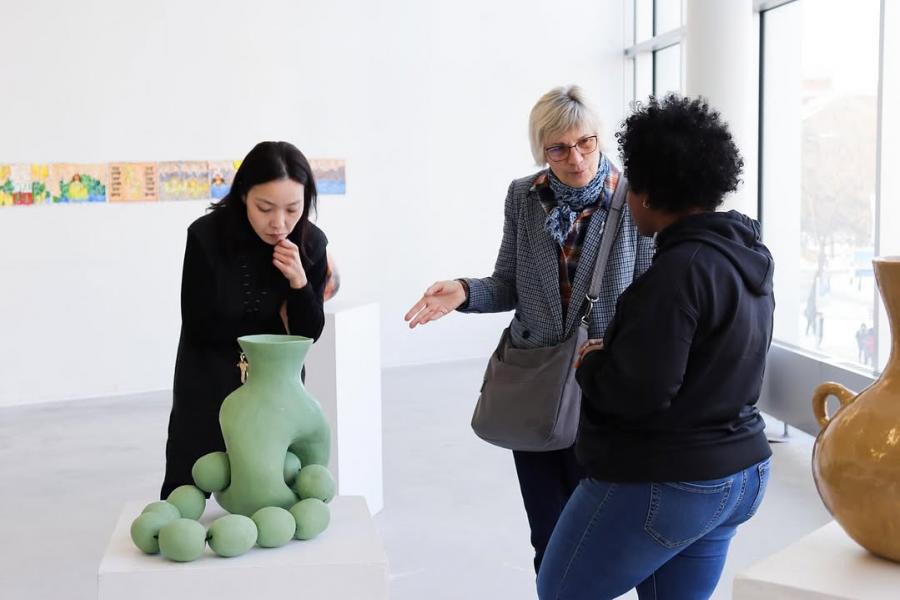 [2:46 PM] Cailyn Harrison Faculty members and student engaged in a discussion around a green ceramic sculpture in a brightly lit gallery space.