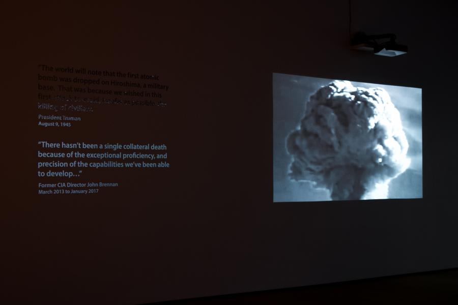A dimly lit gallery space featuring a projection of a mushroom cloud alongside quotes about the atomic bomb.