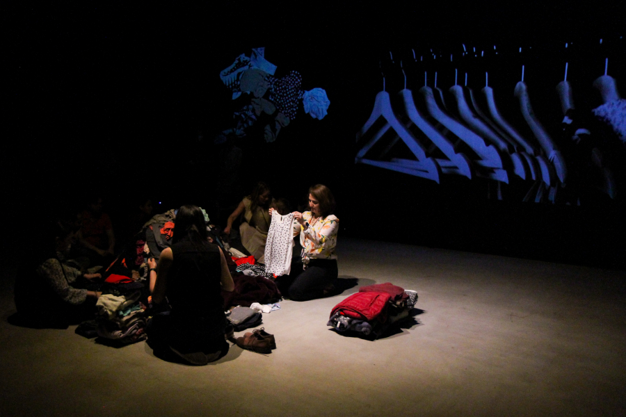 Several participants join the performer, engaging with the pile of clothes, as abstract visuals of hangers and fabrics are projected in the background.